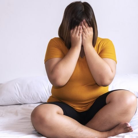 Overweight young woman sitting on white bed while holding hands cover on her face at home. Upset female suffering from extra weight in the bedroom. Obesity unhealthily concept.